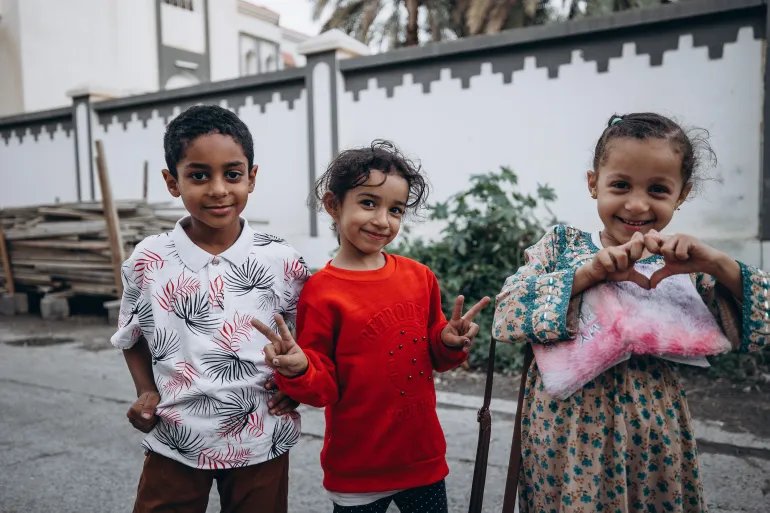 Three smiling children making peace signs in traditional clothing on a street in Nizwa, Oman.
