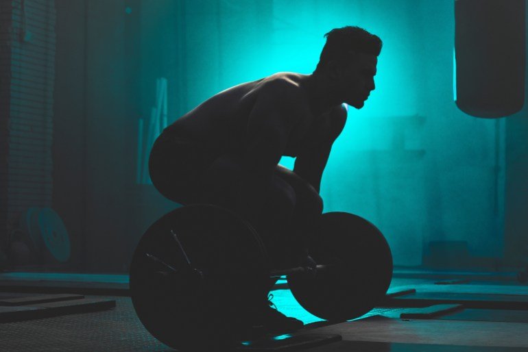 Silhouette, man and weightlifting with barbell for workout, exercise or training in fitness at gym. Active male person or bodybuilder lifting bar for muscle gain, strength or practice at health club