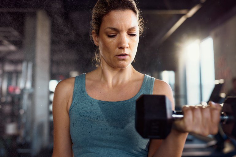Athletic woman making an effort while exercising strength with heavy dumbbell during sports training in a gym.
